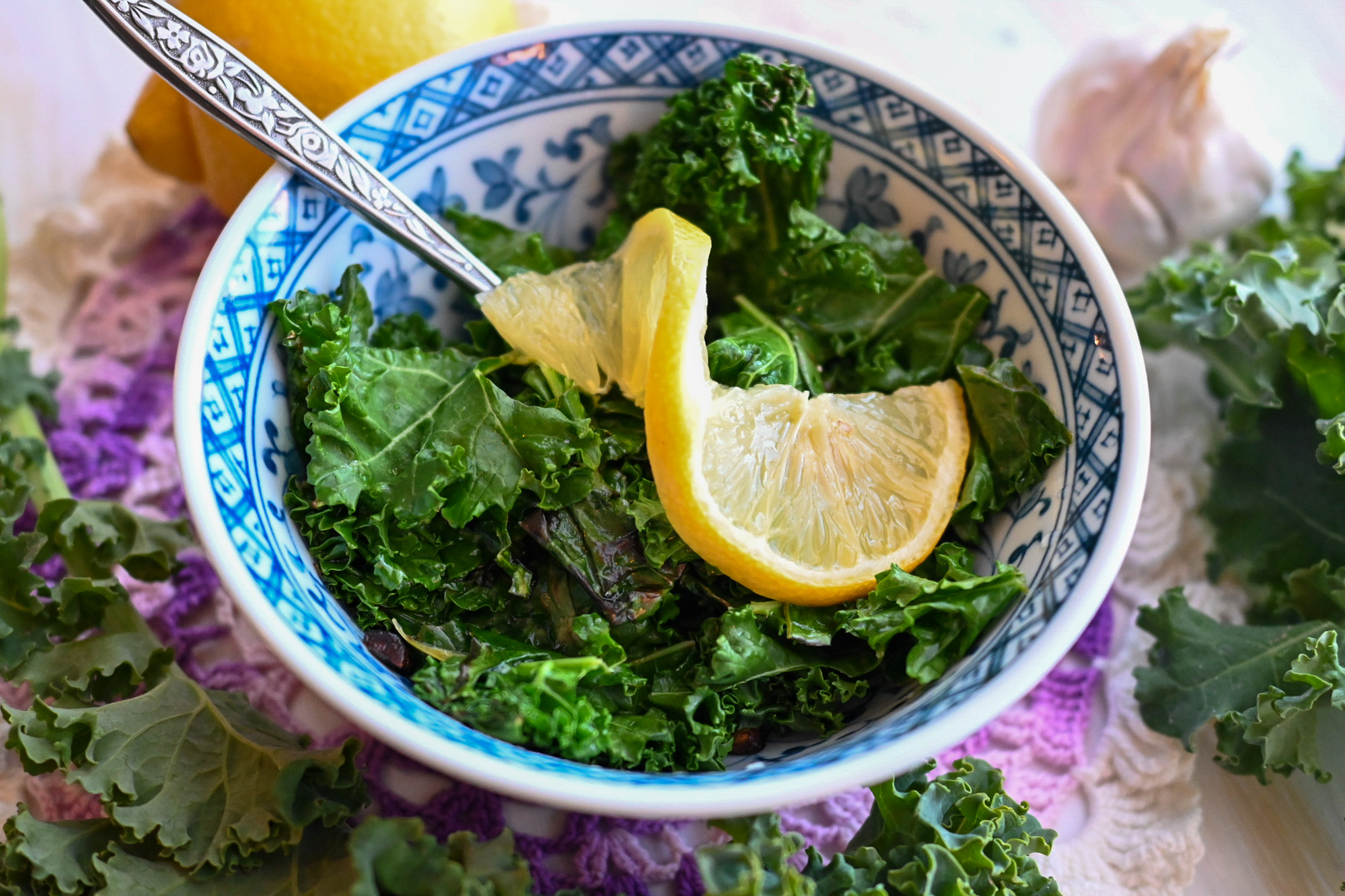 Green curly kale topped with lemon twist in a blue-patterned Japanese ceramic bowl set on purple doily surrounded by a lemon and garlic head.